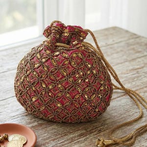 May include: A round, red pouch with intricate gold beadwork and sequins. The drawstring bag features a geometric pattern and a gold cord. The bag is sitting on a wooden surface.