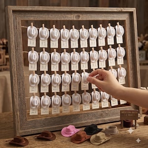 May include: A rustic wooden frame holding rows of twine with miniature white cowboy hats clipped to the strings, each attached to a small paper card. A sign in the corner reads WESTERN HATS - 2 INCH. Loose miniature hats in brown, pink, and black sit below.
