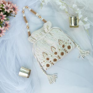May include: A white beaded drawstring bag with a pearl and gold chain handle and tassel. The bag is decorated with pearl and gold floral embroidery.