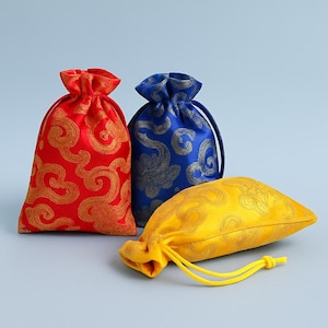 May include: Three decorative drawstring bags in red, blue, and yellow. The red and blue bags are upright, while the yellow bag lies flat. Each bag features a gold swirling pattern. The yellow bag has a yellow drawstring.