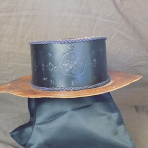 Pode incluir: Um chapéu de couro preto com uma aba larga e uma faixa decorativa. A faixa é feita de um cordão trançado azul escuro e apresenta um design em relevo.