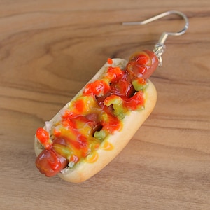 May include: A single, novelty earring shaped like a miniature hot dog. The hot dog is nestled in a bun and topped with ketchup, mustard, and relish. The earring has a silver-colored hook.