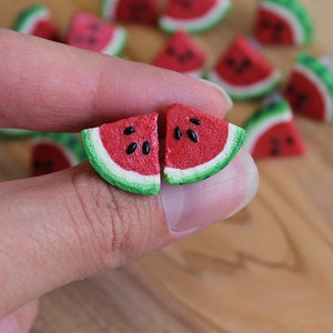 Könnte beinhalten: Zwei kleine Ohrringe in Form von Wassermelonen-Scheiben aus Polymer Clay. Die Ohrringe sind rot mit schwarzen Kernen und einer grünen Rinde.