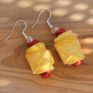 May include: A pair of earrings shaped like pigs in a blanket. The earrings are made of polymer clay and are painted to look like a sausage wrapped in dough. The earrings are hanging from silver hooks.
