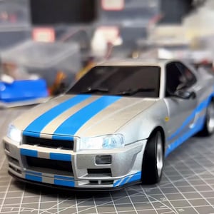 Puede incluir: Modelo de coche plateado con franjas azules. El coche tiene una parrilla delantera detallada, faros y un parabrisas tintado. El modelo es un coupé, con neumáticos y llantas negros. El coche está sobre una superficie gris.