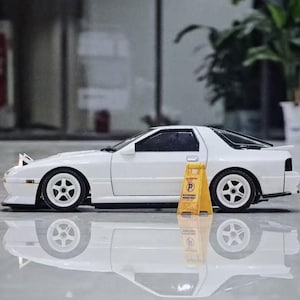 Może przedstawiać: Biały model samochodu sportowego z otwieranymi reflektorami i białymi pięcioramiennymi felgami. Mały żółty plastikowy znak parkingowy z czarnym symbolem P i tekstem znajduje się obok boku samochodu na odblaskowej powierzchni.