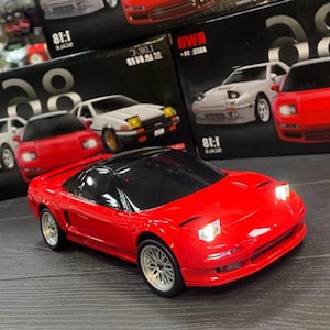 May include: A bright red scale model sports car with a black roof and illuminated pop-up headlights. The vehicle features silver multi-spoke wheels, a low-profile body design, and a dark windshield, presented in front of several product boxes.