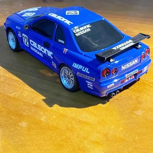 Puede incluir: Un coche de juguete azul con el texto "CALSONIC NISSAN" y el número "12" en el capó y los laterales. El coche tiene un alerón negro y ruedas plateadas. Es un modelo Nissan.