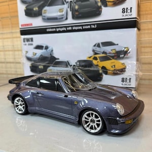 May include: A metallic blue diecast model car with chrome wheels and a rear spoiler. The model is a 1:18 scale replica of a classic sports car. Other model cars are visible in the background.