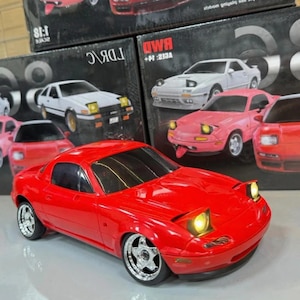 May include: A vibrant red model car with shiny chrome wheels and illuminated headlights. Other model cars are in the background, alongside boxes with car illustrations. The cars are in various colours, including white and pink.