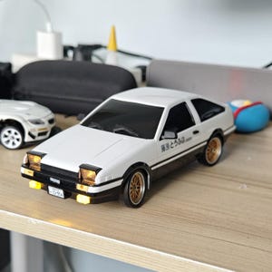 May include: A white and black scale model car featuring pop-up headlights with glowing yellow lights, gold multi-spoke wheels, and Japanese text on the side door. The front bumper displays a license plate with the text 13-954 and the word TRUENO.