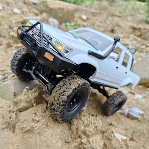 Puede incluir: Un modelo de camioneta pick-up a control remoto de color blanco con protector de parachoques delantero negro, toma de aire tipo snorkel y neumáticos todoterreno grandes cubiertos de barro sobre un terreno rocoso e irregular.
