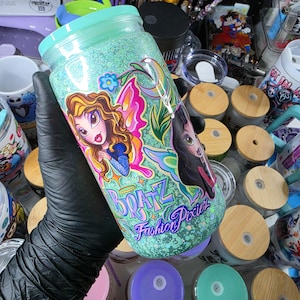 Puede incluir: Un vaso cilíndrico de vidrio con acabado de purpurina verde menta claro y tapa color verde azulado. La superficie muestra dos ilustraciones de personajes tipo hada con alas, acompañadas del texto "BRATZ" y "Fashion Pixiez" en morado.