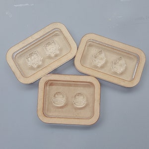 Puede incluir: Tres bandejas rectangulares de madera con insertos de plástico transparente, cada una con dos pequeños moldes transparentes con forma de comida. Los moldes representan una hamburguesa, una porción de pizza y un alimento cuadrado. Las bandejas están dispuestas sobre una superficie gris.