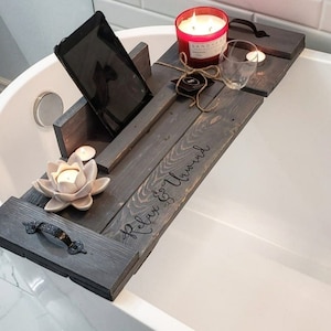 May include: A wooden bath tray with a black finish and the words "Relax & Unwind" inscribed. The tray has a built-in tablet holder, a glass for drinks, and space for candles.