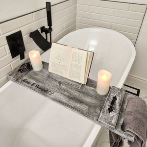 Peut inclure: Un plateau de bain en bois gris avec un porte-livre, deux porte-bougies et un porte-serviette. Le plateau porte le texte "Relax & Unwind" avec un symbole de cœur. Le plateau est dans une baignoire blanche.