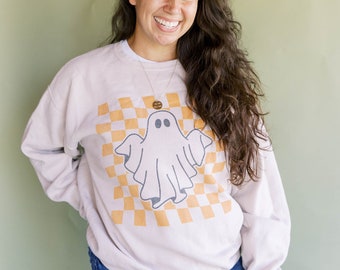 May include: A light beige sweatshirt with a ghost graphic on a yellow and white checkered background. The ghost has two oval eyes and a smiling mouth. The person is wearing blue jeans and a gold necklace.