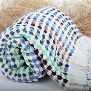 May include: A white, blue, green, and brown patterned Turkish towel with fringe. The towel is rolled up and has a textured surface.