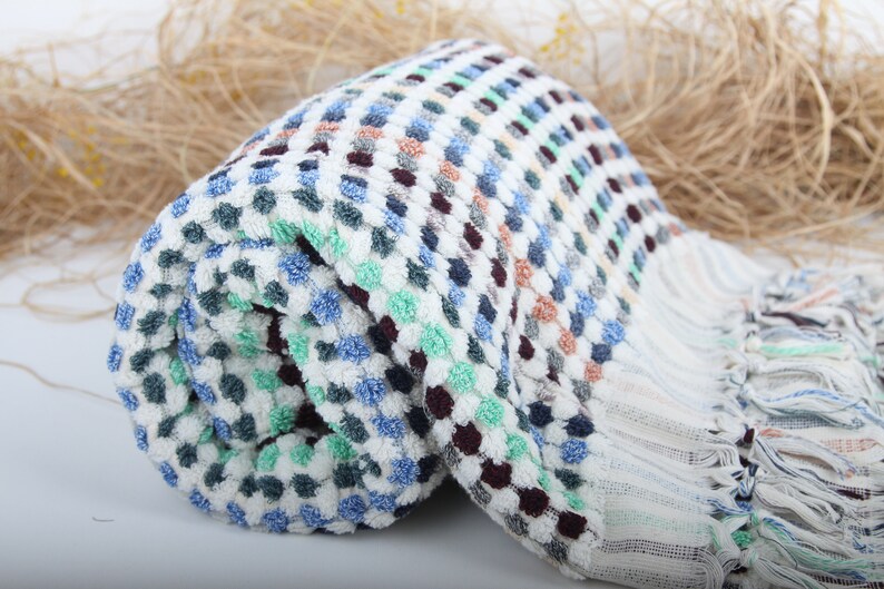 May include: A white, blue, green, and brown patterned Turkish towel with fringe. The towel is rolled up and has a textured surface.