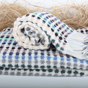 May include: A white bath towel with a checkered pattern in shades of blue, green, and brown. The towel is rolled up and has fringe on the end.