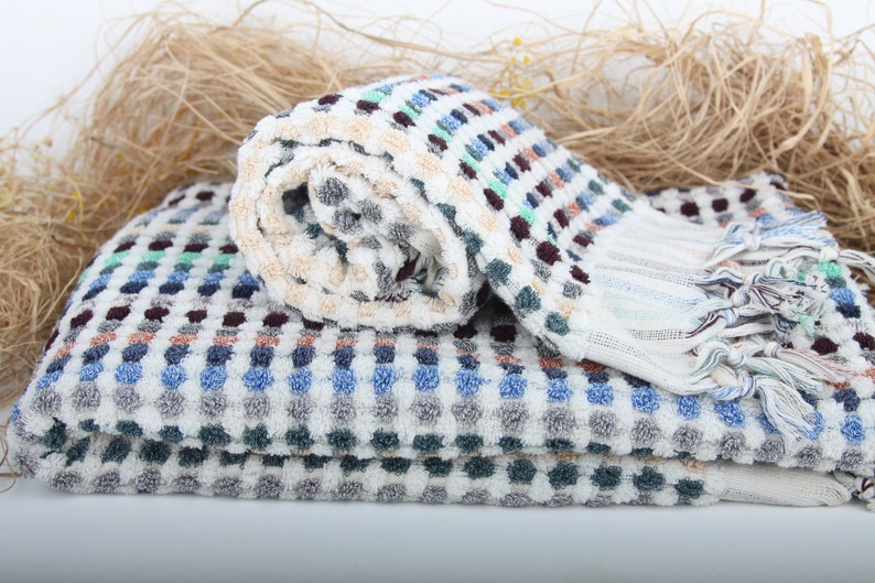 May include: A white bath towel with a checkered pattern in shades of blue, green, and brown. The towel is rolled up and has fringe on the end.