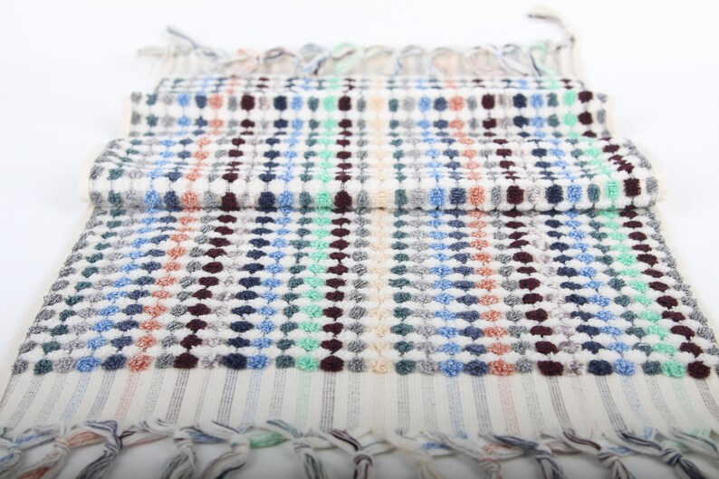 May include: A white hand towel with a colorful geometric pattern of blue, green, red, and brown dots. The towel has a fringe border.