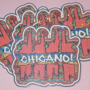 May include: A stack of stickers with a red and white design. The stickers feature the text "CHICANO!" in a stylized font, with a background of roses and a wooden texture. The stickers have a holographic effect.