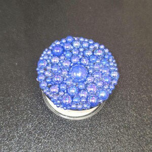 May include: A round, blue container lid covered in iridescent blue beads of varying sizes. The beads create a textured, bubbly surface. The container has a clear, transparent base.