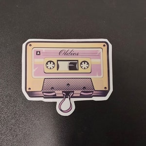 May include: A yellow and pink cassette tape sticker with the word "Oldies" printed on it. The sticker has a string loop attached to the bottom.