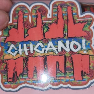 以下が含まれることがあります： 「CHICANO！」の文字が白で、黒い縁取りで描かれたステッカー。カラフルな花柄と幾何学模様の背景。赤、緑、茶色の色合い。ステッカーは手で持たれています。