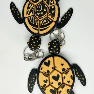 May include: Two turtle-shaped keychains. The turtles have black bodies and limbs with gold-colored segmented shells. One shell features a design, the other has heart shapes. Each keychain has a silver clasp.