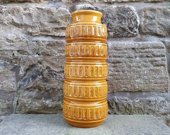 May include: A brown ceramic vase with a geometric pattern. The vase has a cylindrical shape and is decorated with a repeating pattern of squares and rectangles.