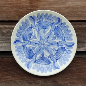 May include: A white ceramic plate with a blue floral design. The plate is circular and features a repeating pattern of stylised flowers.
