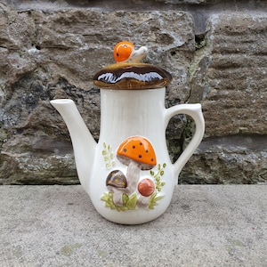 May include: White ceramic teapot with a mushroom lid and mushroom design. The teapot has a brown mushroom lid with an orange top and a brown stem. The teapot has a brown handle and a spout. The teapot is decorated with orange and brown mushrooms and green leaves.