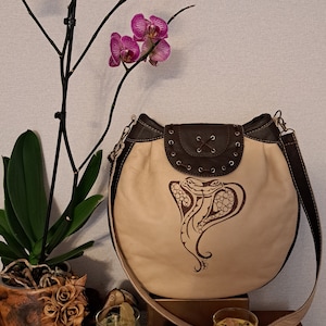 Puede incluir: Un bolso de hombro de cuero beige con una solapa de cuero marrón y un diseño de serpiente. El bolso tiene una correa larga y está decorado con tachuelas metálicas.