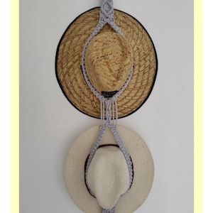 May include: A macrame hat hanger with two straw hats hanging on it. The hanger is made of a light grey cord and is attached to a metal ring. The text "EASY STEPS TO A STYLISH DOUBLE MACRAME HAT HANGER" is at the top of the image. The text "@Krifts_" is at the bottom of the image.