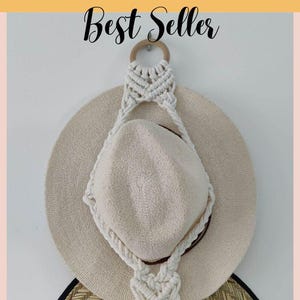 May include: A white macrame hat hanger with a wooden ring at the top. Two straw hats are hanging on the hanger.