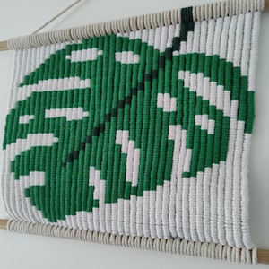 Può includere: Un arazzo da parete in macramè verde e bianco con un motivo a foglia. La foglia è realizzata con filo verde e lo sfondo è in filo bianco.