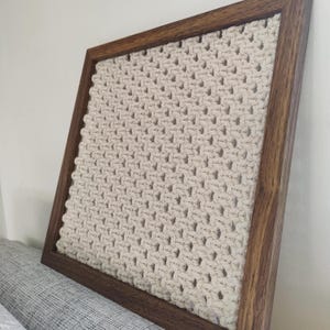 Può includere: Un arazzo da parete in macramè beige con un motivo geometrico, incorniciato in una cornice di legno marrone scuro.