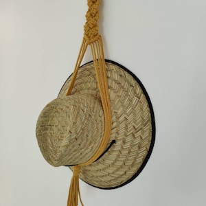 May include: A straw hat with a black trim hangs from a yellow macrame hat holder with a tassel.