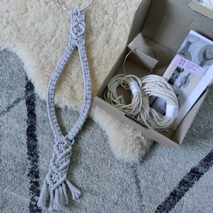 May include: A grey macrame wall hanging with a circular top and tassels at the bottom. The hanging is packaged in a cardboard box with white string and a metal ring.