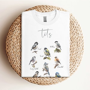 Könnte beinhalten: Weißes T-Shirt mit einer Grafik verschiedener Meisenarten, darunter eine Kohlmeise, eine Blaumeise, eine Schwanzmeise, eine Haubenmeise, eine Bartmeise und eine Sumpfmeise. Der Text "Tits" ist über den Meisen gedruckt.