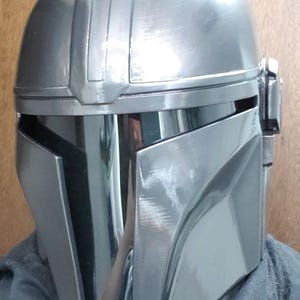 K&ouml;nnte beinhalten: Ein metallisch silberner Integralhelm mit einer glatten, abgerundeten Kuppel, einem zentralen vertikalen Grat und einem dunklen, reflektierenden T-f&ouml;rmigen Visier. Der Helm hat eine polierte Oberfl&auml;che mit sichtbaren Pinselstrichen.