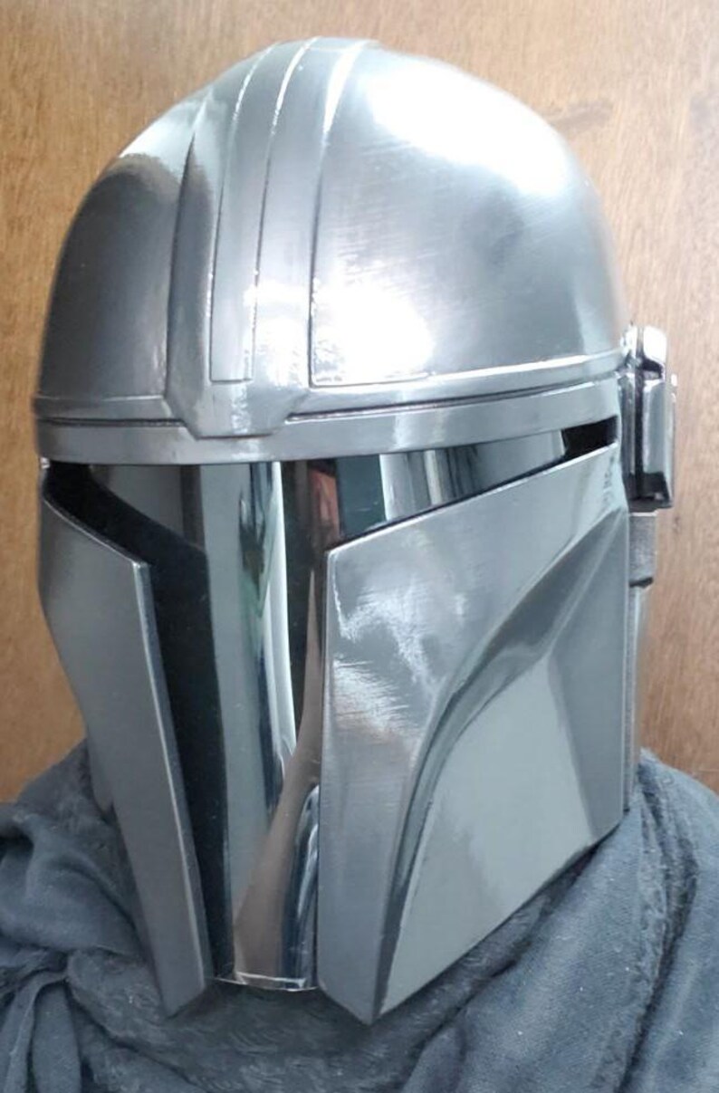 K&ouml;nnte beinhalten: Ein metallisch silberner Integralhelm mit einer glatten, abgerundeten Kuppel, einem zentralen vertikalen Grat und einem dunklen, reflektierenden T-f&ouml;rmigen Visier. Der Helm hat eine polierte Oberfl&auml;che mit sichtbaren Pinselstrichen.
