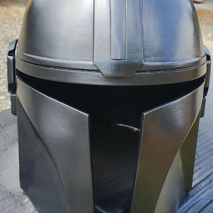 K&ouml;nnte beinhalten: Ein mattschwarzer Helm mit einer glatten, abgerundeten Kuppel, einem zentralen vertikalen Grat und einer breiten, horizontalen T-f&ouml;rmigen Visier&ouml;ffnung. Der Helm hat ein strukturiertes, kantiges Frontdesign mit seitlichen Ohrenkappen.