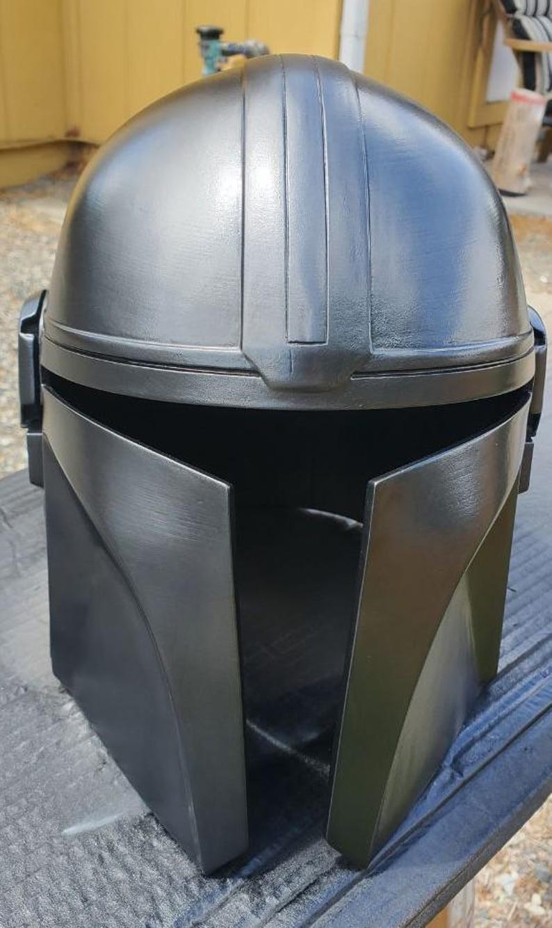 K&ouml;nnte beinhalten: Ein mattschwarzer Helm mit einer glatten, abgerundeten Kuppel, einem zentralen vertikalen Grat und einer breiten, horizontalen T-f&ouml;rmigen Visier&ouml;ffnung. Der Helm hat ein strukturiertes, kantiges Frontdesign mit seitlichen Ohrenkappen.
