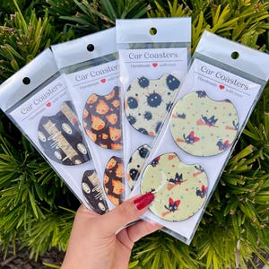 May include: Four sets of car coasters with different designs. Each set has a white background with a black and yellow pattern. The designs include a black cat, a black cat with a broom, a black cat with a witch's hat, and a black cat with a pumpkin. The text on the packaging reads "Car Coasters Handmade with love."