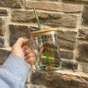 May include: A glass mug with a bamboo lid and a green straw. The mug has a graphic of the entrance to a hobbit hole with the text "There and Back Again A Hobbits Tale".