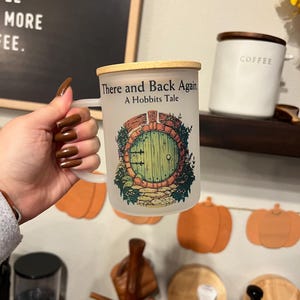 May include: White ceramic mug with a wooden lid. The mug has a graphic of a green door with a stone path leading to it. The text "There and Back Again A Hobbits Tale" is printed on the mug.