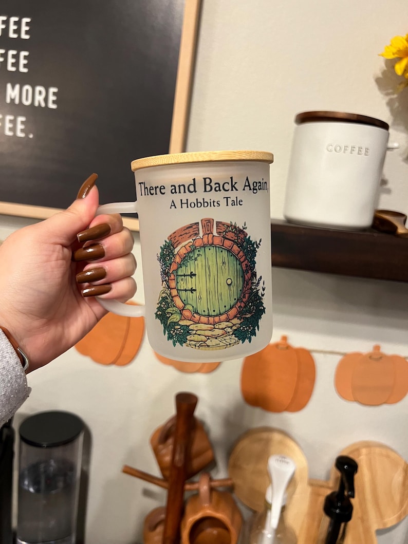 May include: White ceramic mug with a wooden lid. The mug has a graphic of a green door with a stone path leading to it. The text "There and Back Again A Hobbits Tale" is printed on the mug.
